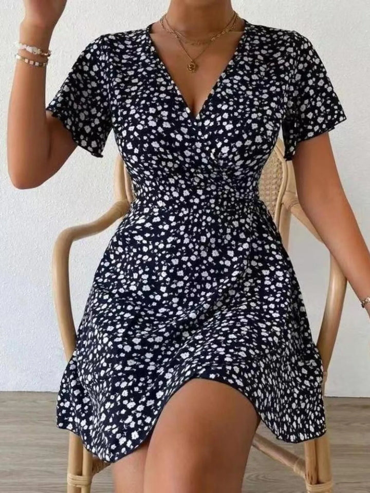 Woman wearing a black floral dress sitting on a chair against a white wall.