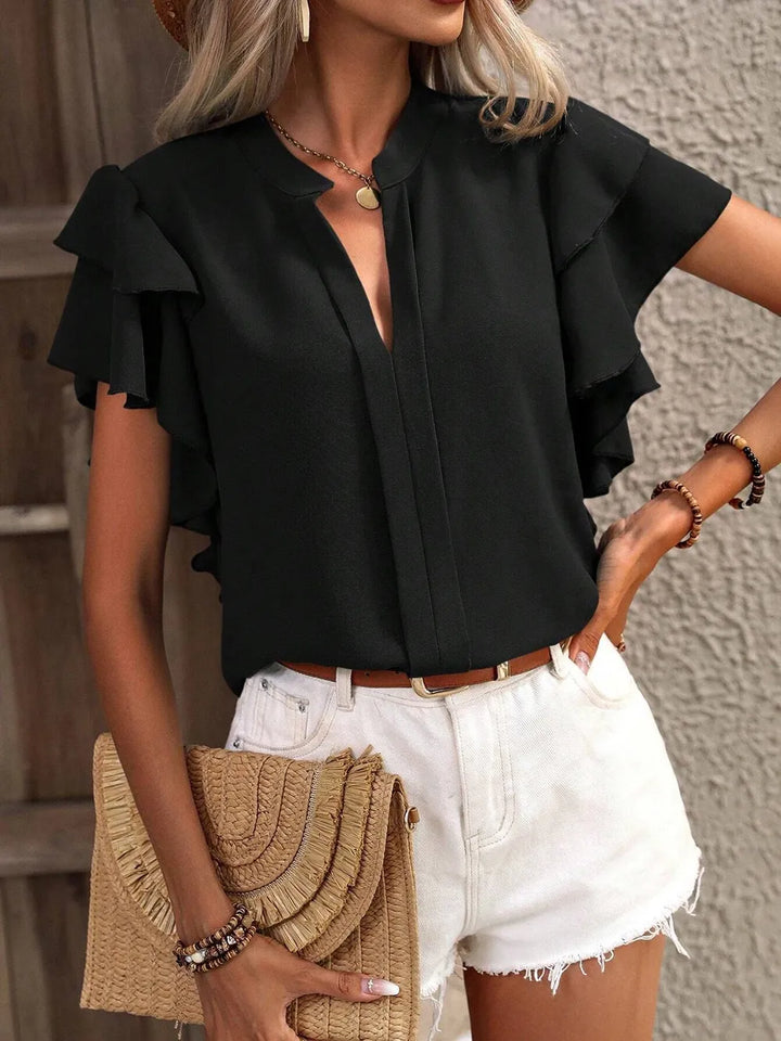Woman wearing a black ruffled blouse and white shorts, holding a beige clutch.