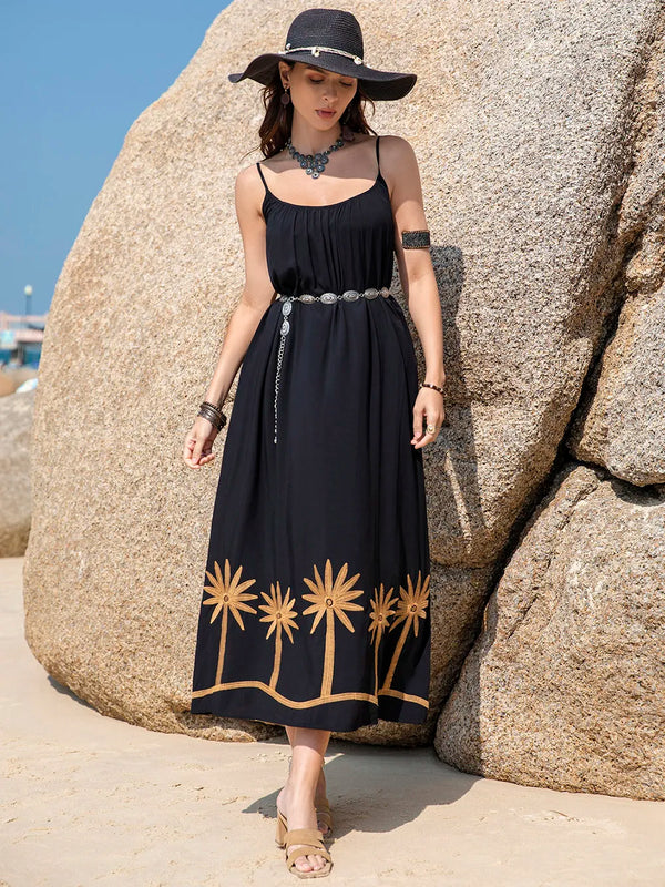 Woman in a black dress with gold embroidery standing against large rocks on a beach.