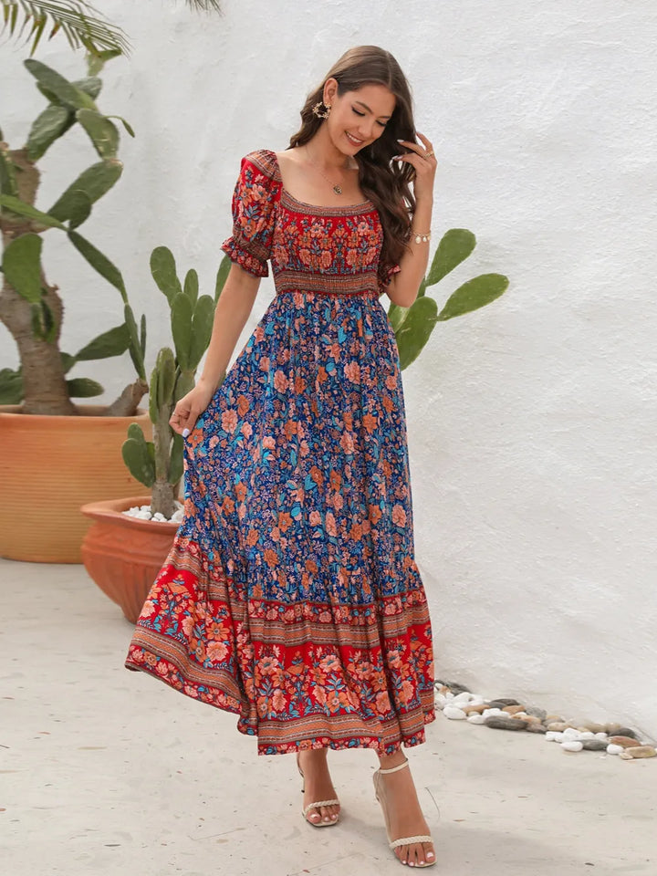 Woman wearing a colorful floral midi dress standing in front of a white wall with plants.