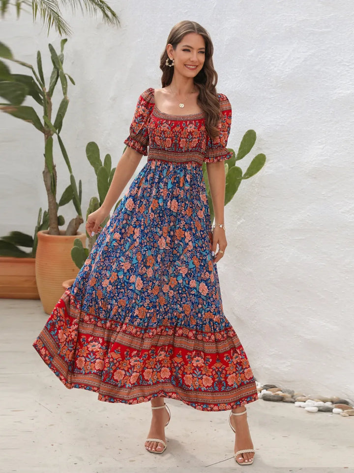 Woman wearing a colorful midi dress with a pattern of flowers and leaves, standing against a white wall with plants.
