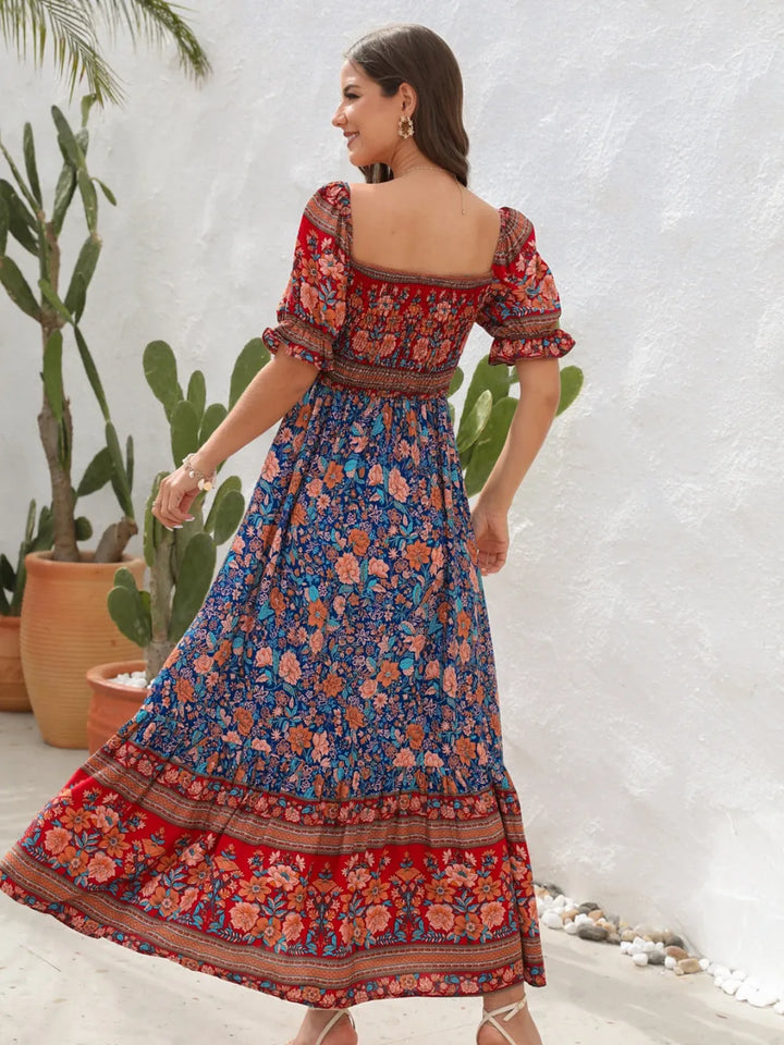 Woman wearing a colorful floral midi dress with a white textured wall and plants in the background