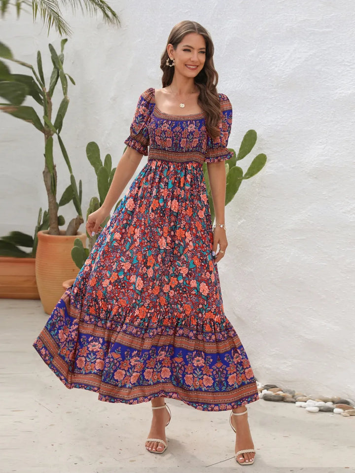 Woman wearing a colorful floral midi dress standing in front of a white wall with plants.