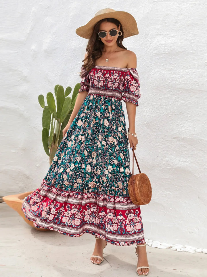 Woman wearing a floral off-shoulder midi dress with a wide-brimmed hat and sunglasses, standing against a white wall with a cactus plant.