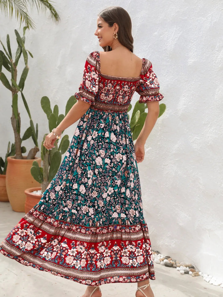 Woman wearing a colorful floral midi dress with a white textured wall and plants in the background