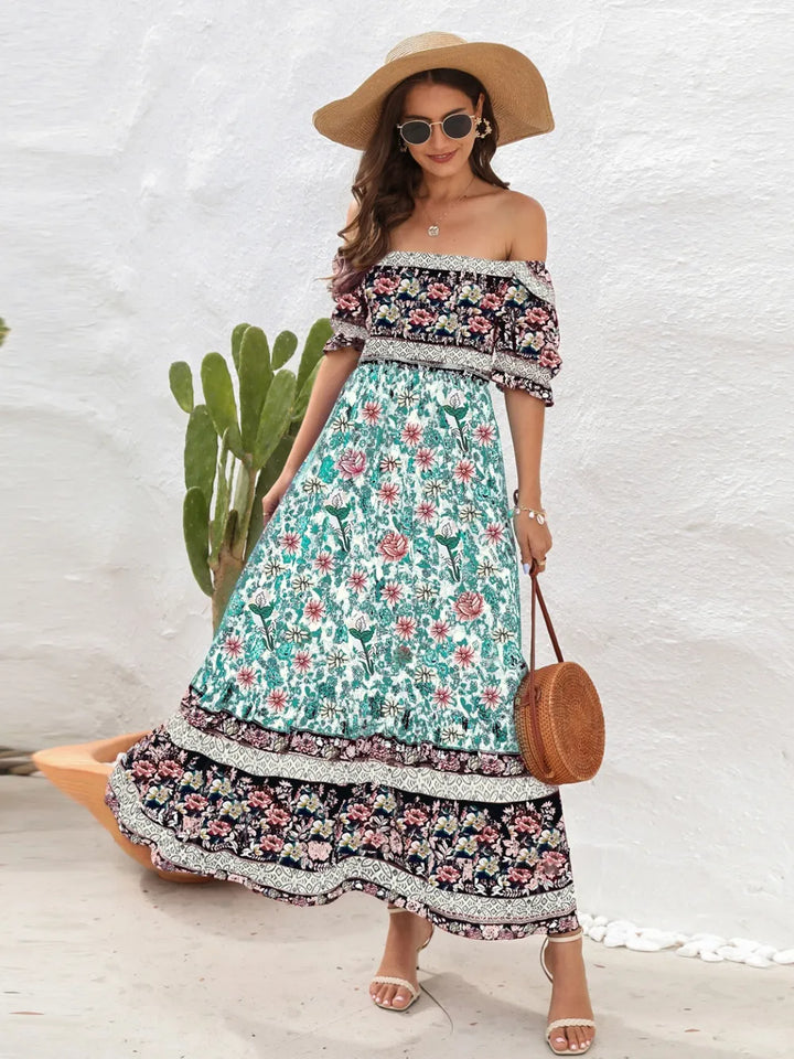 Woman wearing a floral off-shoulder midi dress with a straw hat and sunglasses, standing against a white wall.