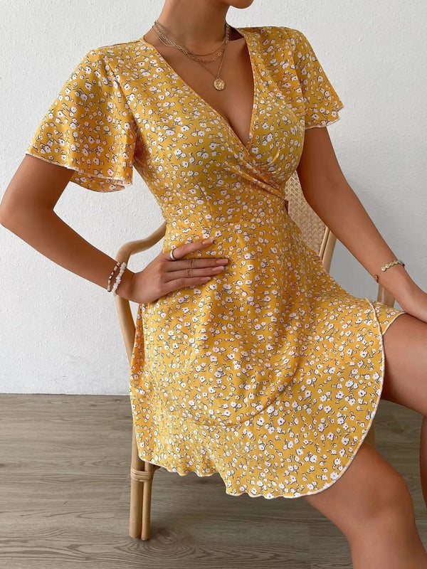 Woman wearing a yellow floral dress sitting on a wooden chair against a white wall.