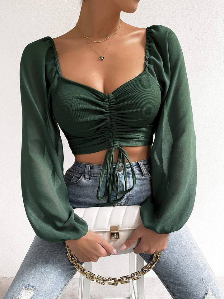 Person wearing a green ruched cropped top with long sleeves and light blue jeans, holding a white clutch.