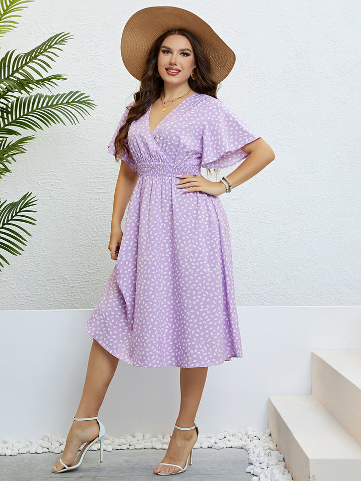 Woman wearing a lavender printed midi dress with a wide-brimmed hat against a white wall.