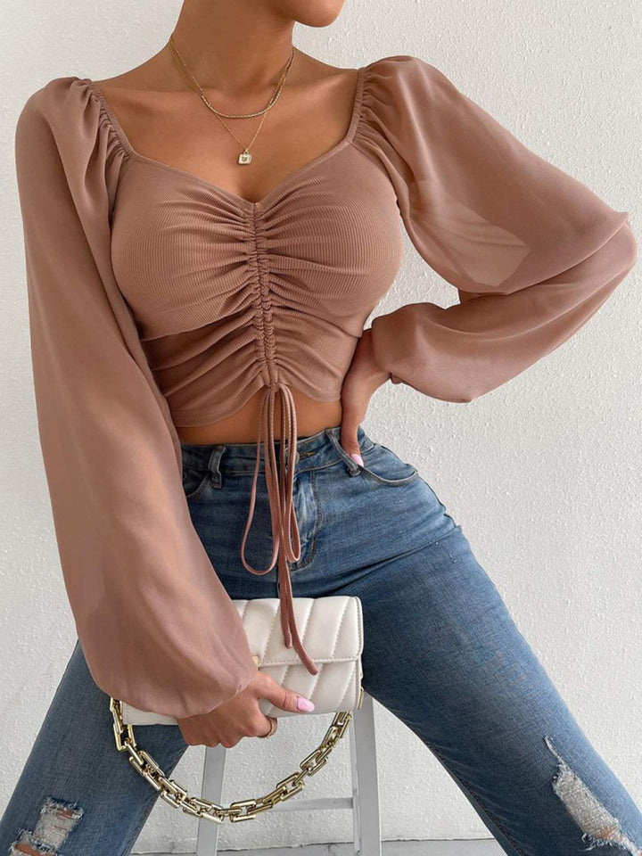 Woman wearing a beige ruched cropped top and blue jeans, holding a beige handbag.