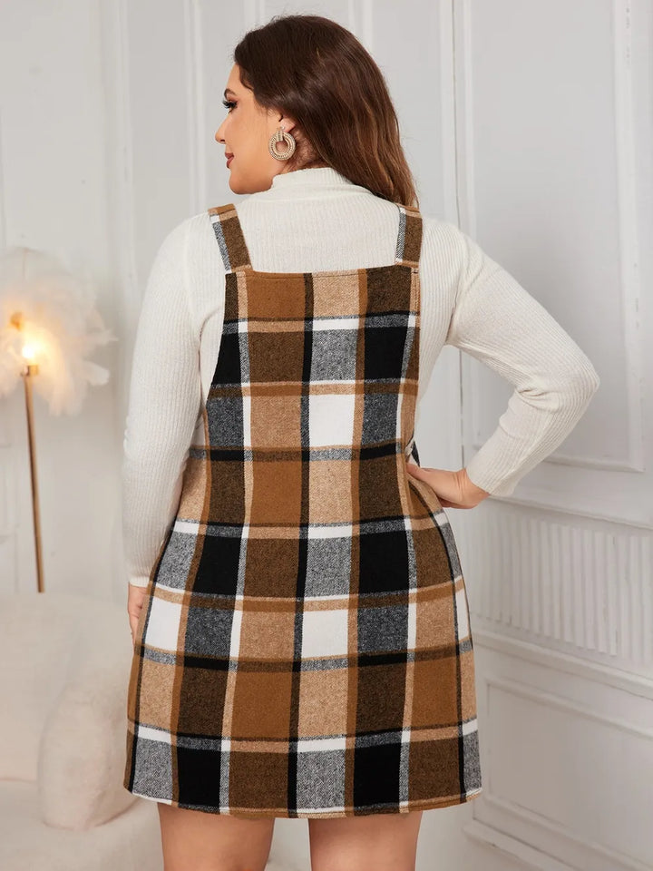 Woman wearing a plaid pinafore dress with a white sweater underneath, standing against a white wall.