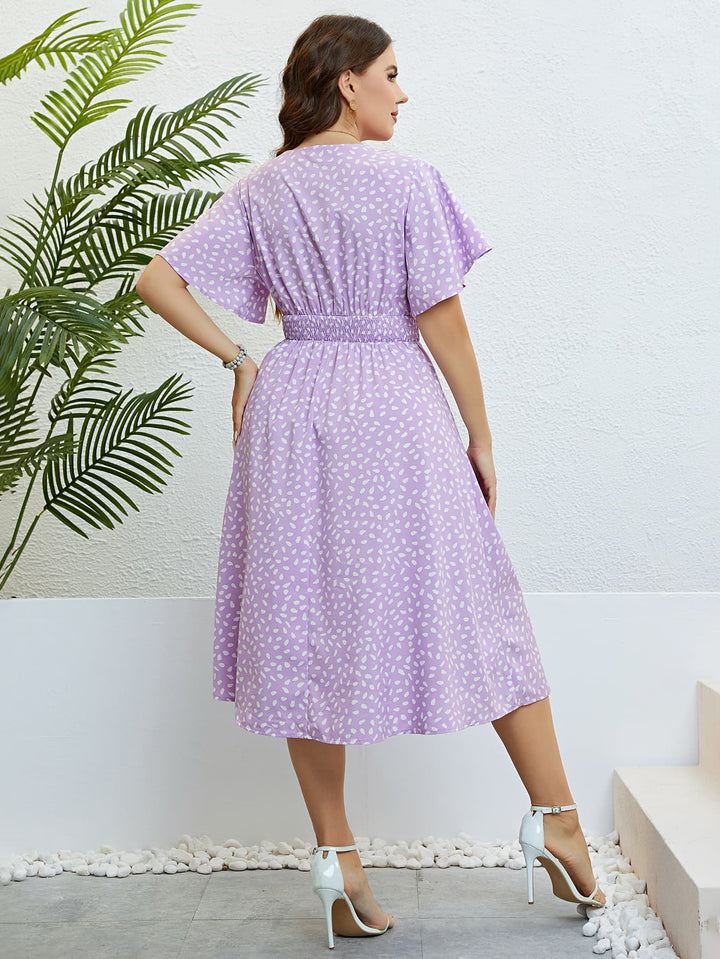 Woman wearing a lavender printed midi dress standing against a white wall with a plant in the background.