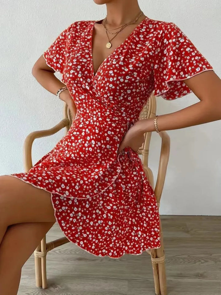 Woman wearing a red floral dress sitting on a chair against a white wall.