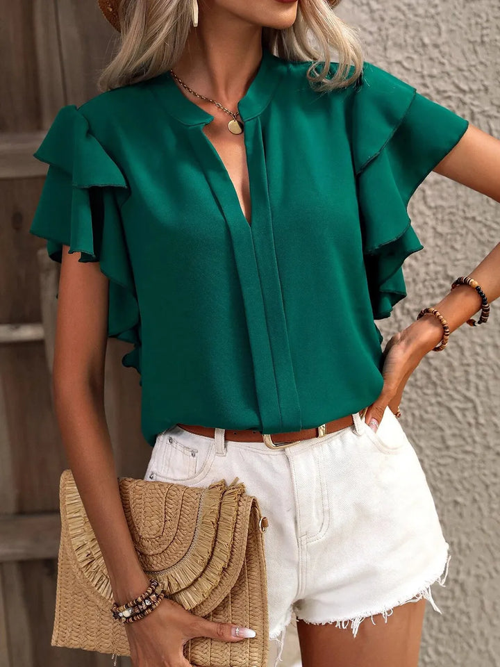 Woman wearing a green blouse with ruffled sleeves and white shorts, holding a straw clutch.