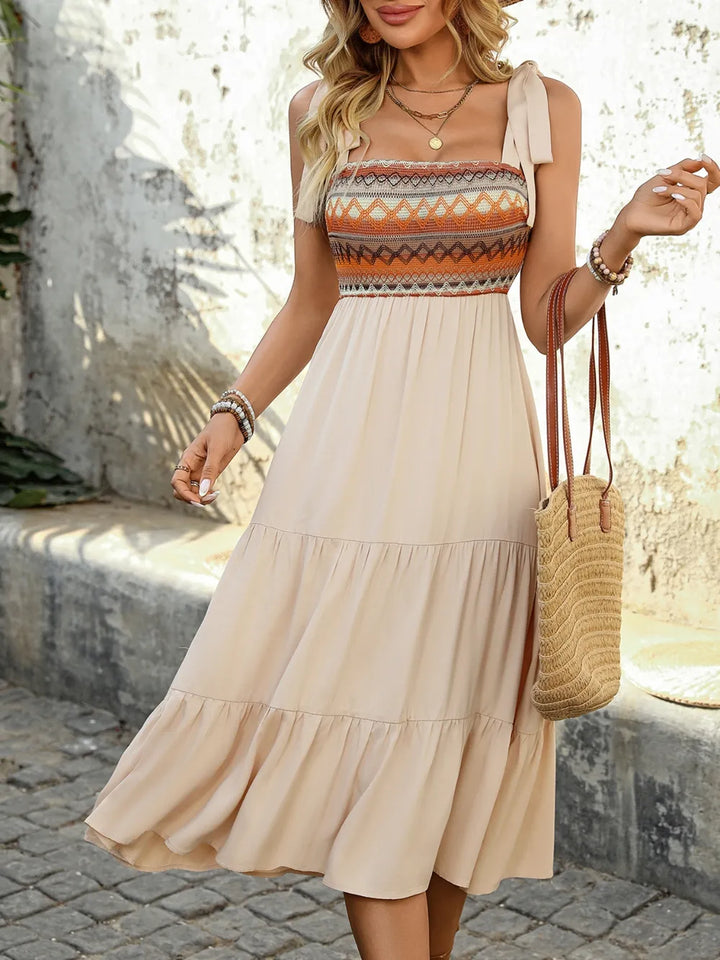 Woman wearing a beige dress with a patterned top, holding a straw bag outdoors.