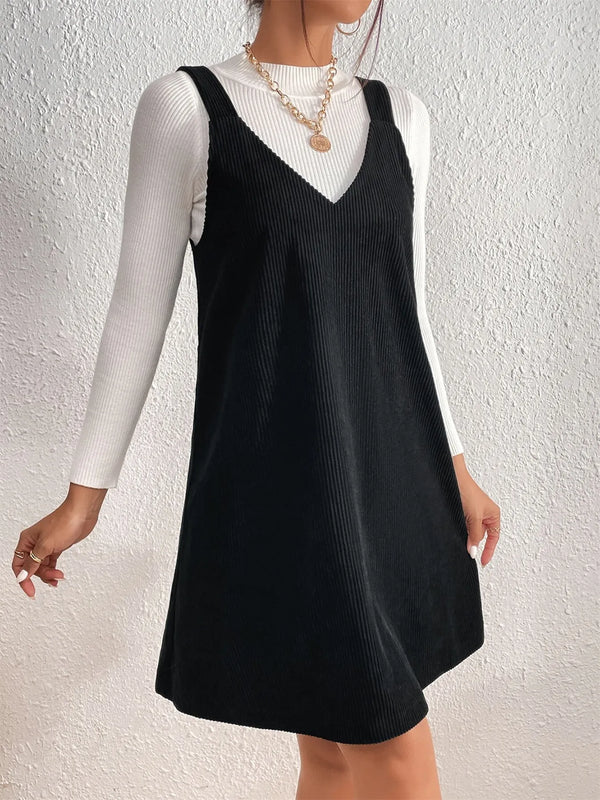 Person wearing a black pinafore dress over a white long-sleeve shirt against a light background