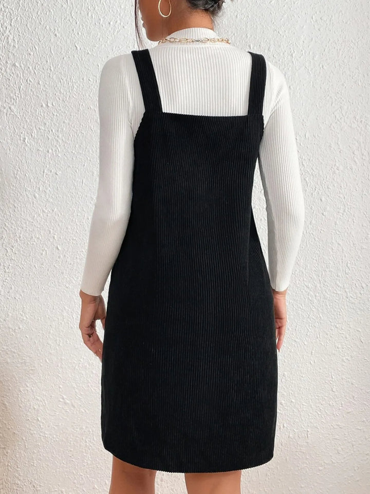 Person wearing a black pinafore dress over a white long-sleeve shirt against a white background